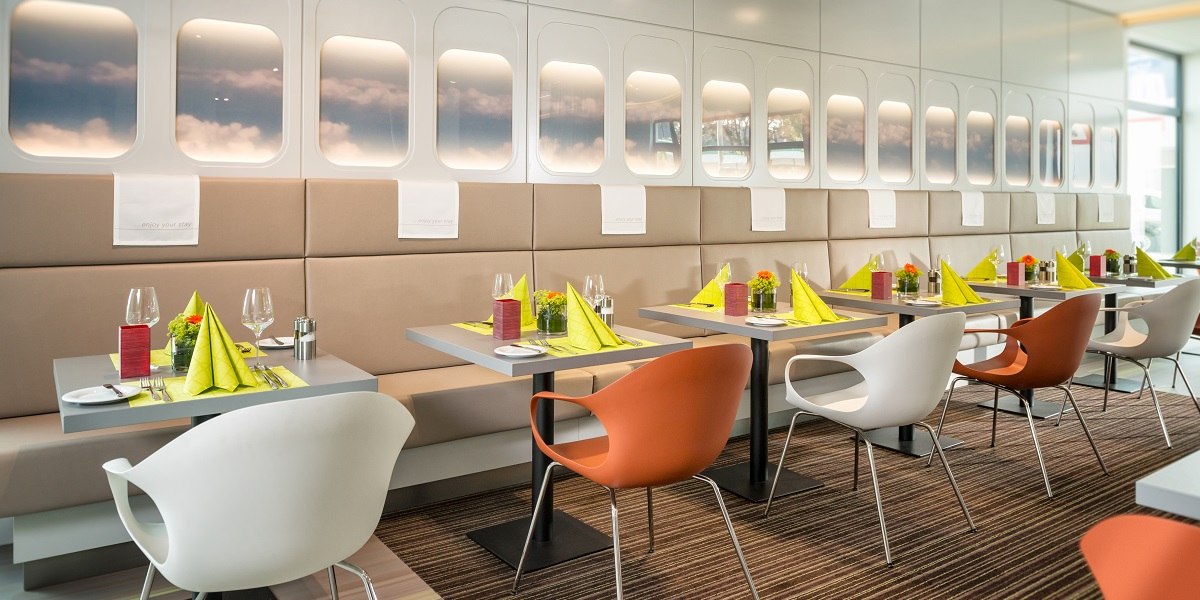 Modernes Restaurant mit Flugzeugfenster-Dekor, gedeckte Tische mit grünen Servietten und bunten Stühlen., © Essential by Dorint
