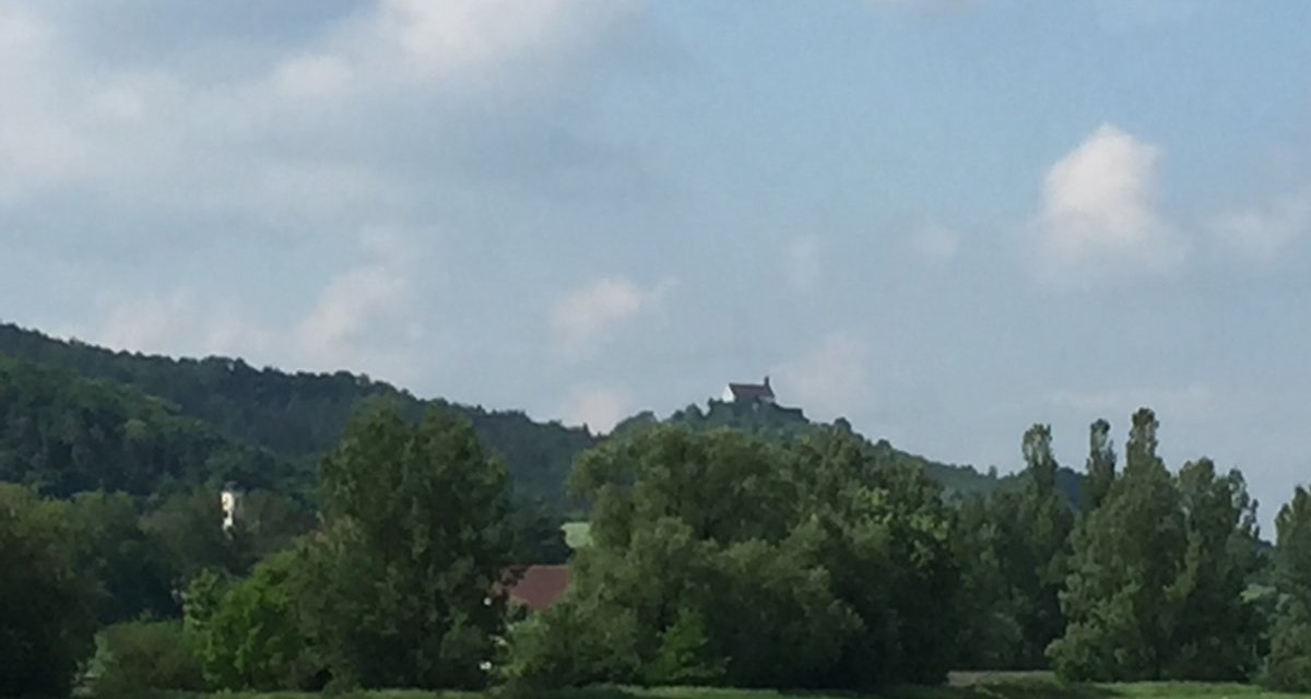 Blick auf eine grüne Landschaft mit einem Hügel im Hintergrund, auf dem eine Kapelle steht. Der Himmel ist leicht bewölkt., © www.pro-cycl.de Blick auf eine grüne Landschaft mit einem Hügel im Hintergrund, auf dem eine Kapelle steht. Der Himmel ist leicht bewölkt., © www.pro-cycl.de