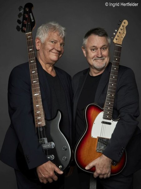 Zwei M&auml;nner in dunklen Anz&uuml;gen posieren mit Gitarren vor einem grauen Hintergrund und l&auml;cheln in die Kamera., &copy; Stadthalle Leonberg