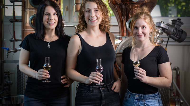 Drei Frauen in schwarzer Kleidung stehen in einer Brauerei und halten Gl&auml;ser mit Getr&auml;nken. Im Hintergrund sind Brauereiger&auml;te zu sehen., &copy; Brau- & Brennwerkstatt