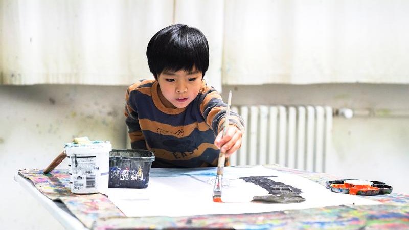 Ein Kind malt konzentriert mit einem Pinsel auf Papier. Der Tisch ist mit Farben und Malutensilien bedeckt. Im Hintergrund ist ein Heizk&ouml;rper zu sehen., &copy; Kunstschule Labyrinth