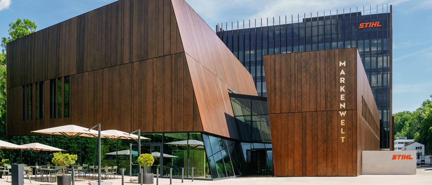 Moderne Gebäude der STIHL Markenwelt mit Holzverkleidung und Glasfronten, umgeben von Bäumen. Sonnenschirme und Sitzgelegenheiten im Vordergrund., © STIHL Markenwelt