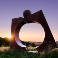 Eine gro&szlig;e Metallskulptur steht auf einem Feld, w&auml;hrend die Sonne am Horizont untergeht. Menschen spazieren und radeln im Hintergrund., &copy; Andreas Sporn