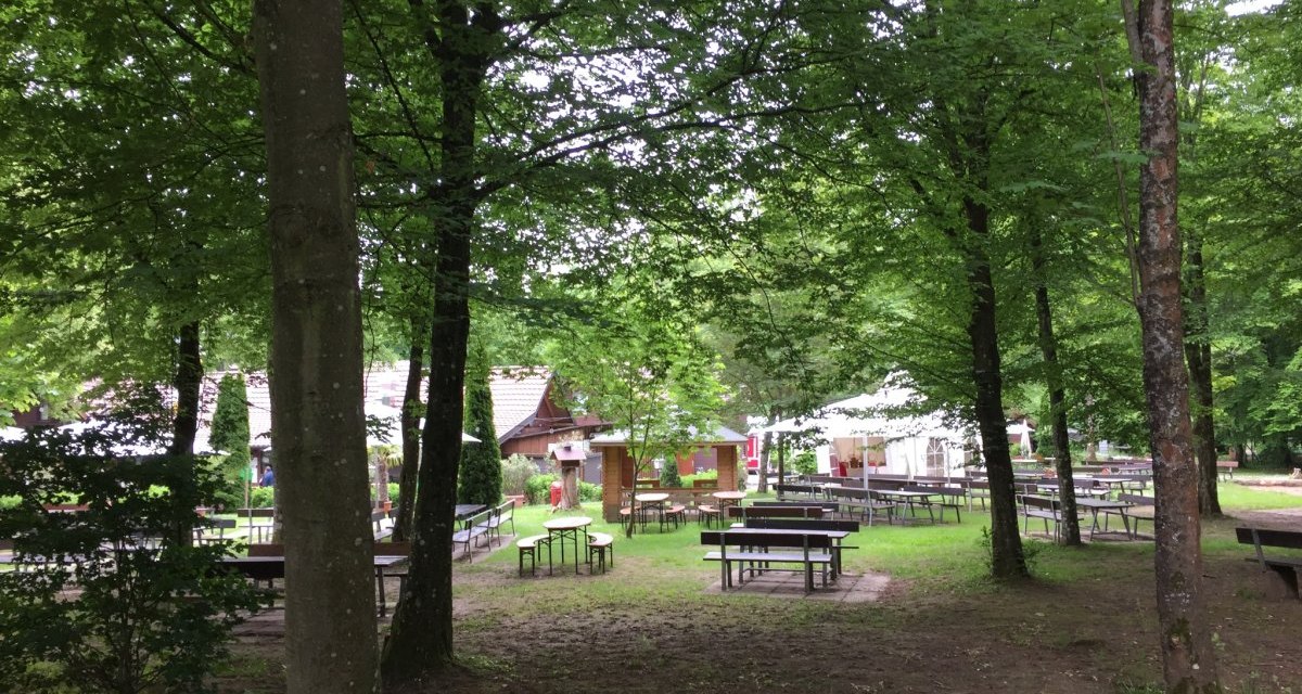 Ein idyllischer Biergarten im Wald mit leeren Holztischen und B&auml;nken, umgeben von gr&uuml;nen B&auml;umen und einem kleinen Geb&auml;ude im Hintergrund., &copy; www.pro-cycl.de