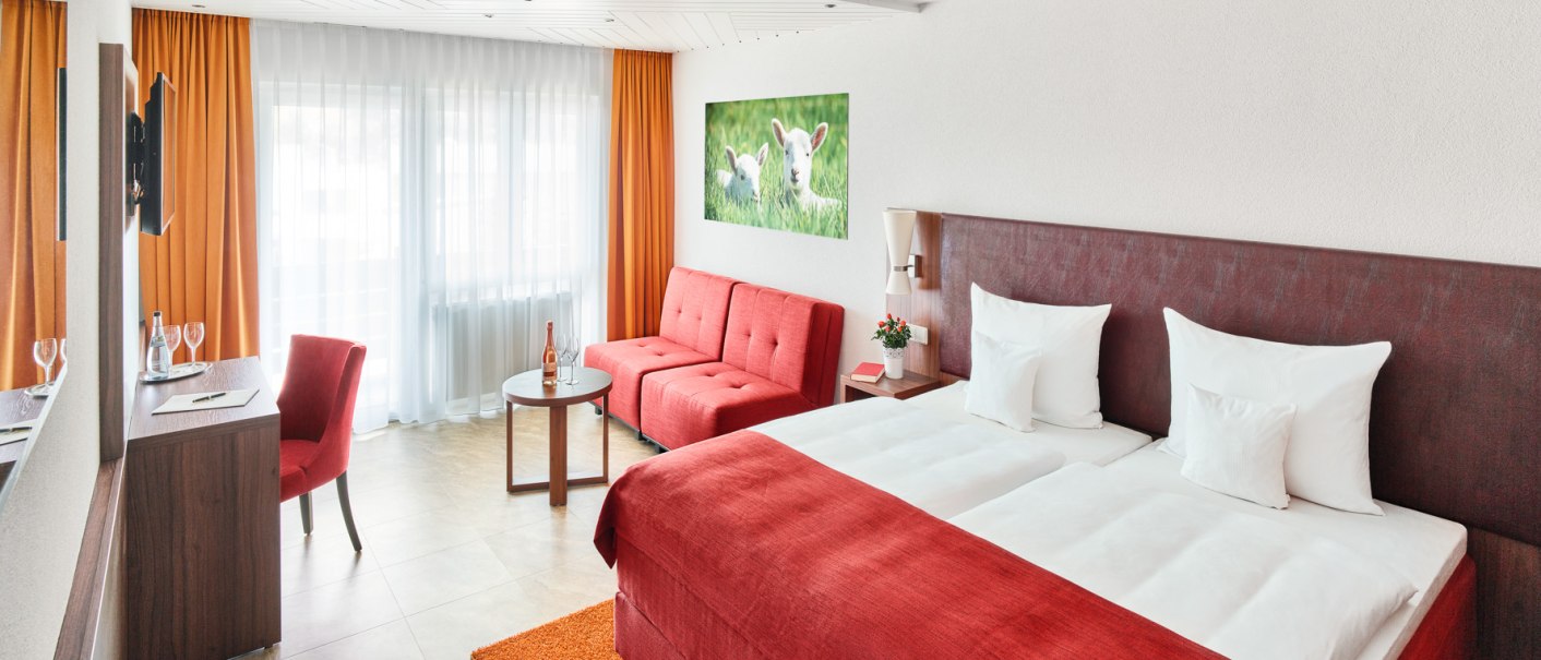 Modernes Hotelzimmer mit Doppelbett, rotem Sofa, Schreibtisch und großem Fenster mit Vorhängen. Ein Bild von Lämmern hängt an der Wand., © Hotel Restaurant Lamm