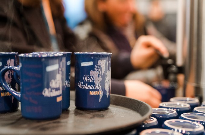 Blaue Tassen mit 'Welt Weihnachts Markt' stehen auf einem Tablett. Im Hintergrund schenken Menschen Glühwein ein, was eine festliche Atmosphäre schafft., © Cool-Tours StattReisen Blaue Tassen mit 'Welt Weihnachts Markt' stehen auf einem Tablett. Im Hintergrund schenken Menschen Glühwein ein, was eine festliche Atmosphäre schafft., © Cool-Tours StattReisen