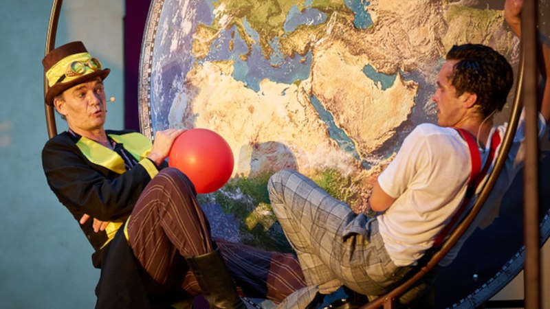 Zwei M&auml;nner in Kost&uuml;men sitzen vor einer gro&szlig;en Weltkarte. Einer tr&auml;gt einen Zylinder und h&auml;lt einen roten Ballon., &copy; Kultur- und Sportamt Stadt Bietigheim-Bissingen