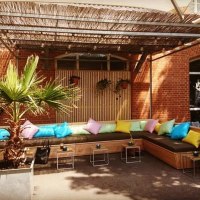Terrasse mit bunten Kissen auf einer Eckbank, Palmen in Töpfen und einem Holzdach. Ziegelwand im Hintergrund., © Blauer Engel GmbH Terrasse mit bunten Kissen auf einer Eckbank, Palmen in Töpfen und einem Holzdach. Ziegelwand im Hintergrund., © Blauer Engel GmbH
