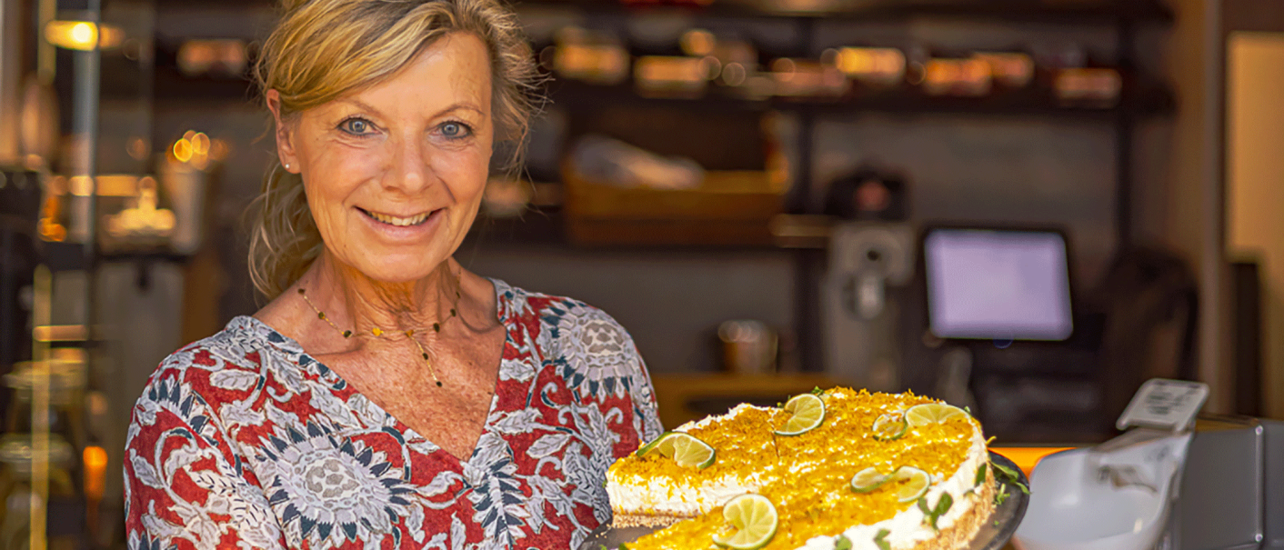 Eine lächelnde Frau in einem Café hält eine Torte mit Limettenscheiben und gelbem Belag. Im Hintergrund sind Regale und ein Computer zu sehen., © Ama Deli Eine lächelnde Frau in einem Café hält eine Torte mit Limettenscheiben und gelbem Belag. Im Hintergrund sind Regale und ein Computer zu sehen., © Ama Deli