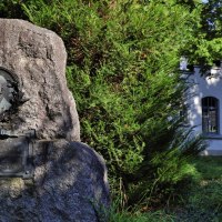Gedenkstein für Gottlieb Daimler in einem grünen Park, rechts ein weißes Gebäude mit Glasfenstern., © Stuttgart-Marketing GmbH Achim Mende