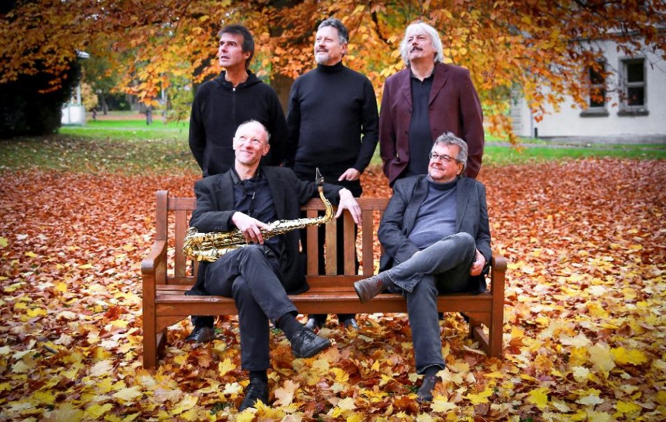 F&uuml;nf M&auml;nner in herbstlicher Umgebung, zwei sitzen auf einer Bank, einer h&auml;lt ein Saxophon. Bunte Bl&auml;tter bedecken den Boden., &copy; Foto Doris Bredow