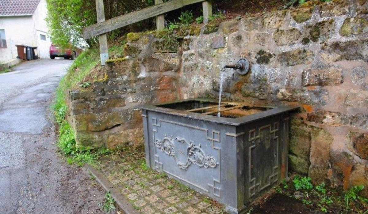 Ein historischer Brunnen mit fließendem Wasser, eingebaut in eine Steinmauer. Der Brunnen hat dekorative Verzierungen., © Bürgerausschuss WHSO