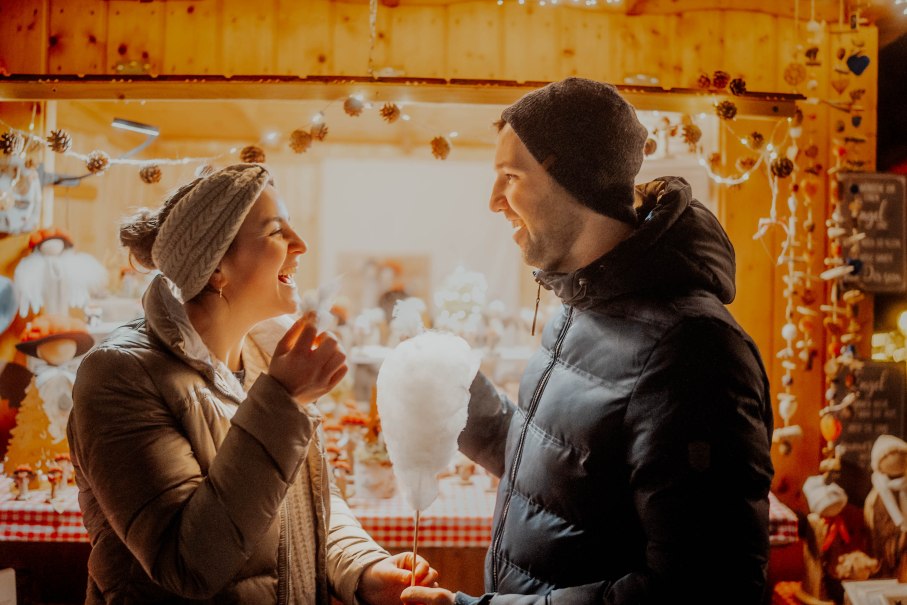 Ein Paar lacht und genießt Zuckerwatte vor einem festlich dekorierten Stand auf einem Weihnachtsmarkt., © Nik Ebert