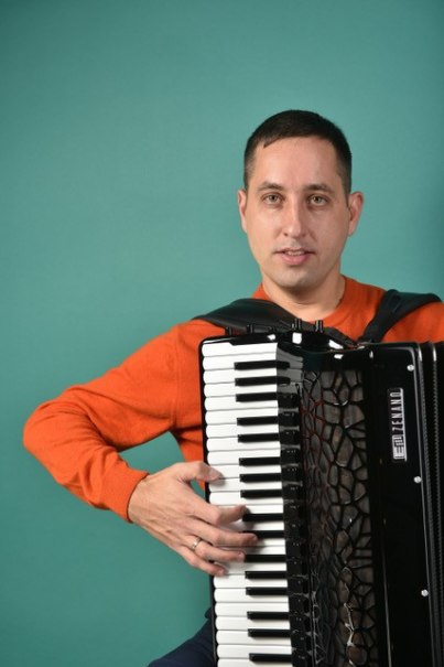 Ein Mann in einem orangefarbenen Pullover spielt Akkordeon vor einem türkisfarbenen Hintergrund. Er schaut in die Kamera., © Roman Dotsenko Ein Mann in einem orangefarbenen Pullover spielt Akkordeon vor einem türkisfarbenen Hintergrund. Er schaut in die Kamera., © Roman Dotsenko