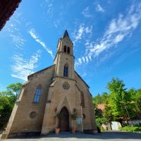 Die Evangelische Schlosskirche Remseck-Hochberg vor blauem Himmel mit dekorativen Wolken. Der Kirchturm ragt in die Höhe, umgeben von Bäumen.