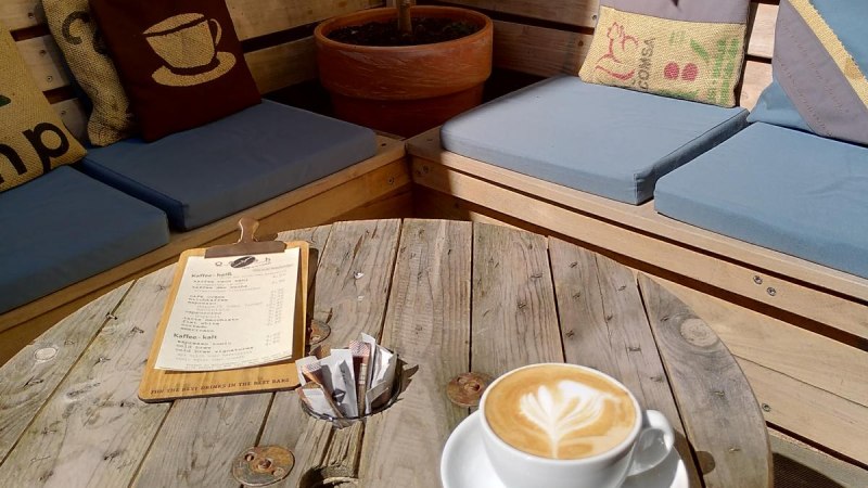 Gemütlicher Außenbereich mit Holzmöbeln, Kissen und Pflanzen. Auf dem Tisch steht eine Tasse Cappuccino und eine Speisekarte., © Matthias Gohl