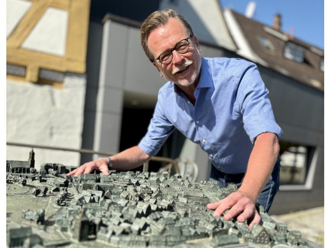 Ein Mann in einem blauen Hemd beugt sich l&auml;chelnd &uuml;ber ein detailliertes Stadtmodell. Im Hintergrund sind Geb&auml;ude und blauer Himmel zu sehen., &copy; WTM GmbH Waiblingen