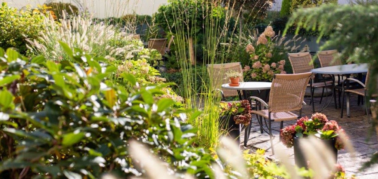 Ein sonniger Garten mit Tischen und Stühlen, umgeben von üppigen Pflanzen und Blumen. Im Hintergrund sind Gebäude zu sehen., © Kronen Hotel GmbH