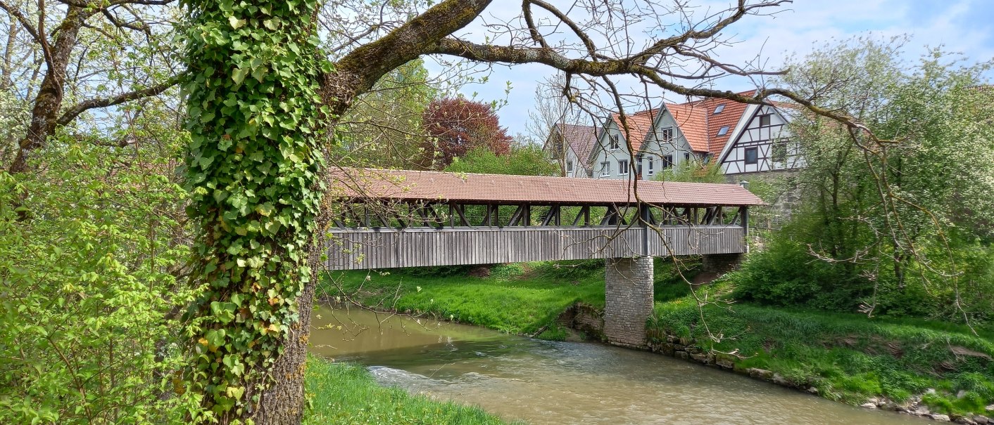 Fluss mit &uuml;berdachter Holzbr&uuml;cke, am Ufer B&uuml;sche und B&auml;ume, auf der rechten Bildseite sind die wei&szlig;en Giebelseiten und rotbraunen Dachfl&auml;chen von vier H&auml;usern hinter der Holzbr&uuml;cke zu erkennen, &copy; Petra Natzkowski