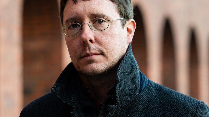 Ein Mann mit Brille und dunklem Mantel steht vor einer Backsteinmauer mit B&ouml;gen. Er blickt ernst in die Kamera., &copy; Kulturforum Schorndorf e. V.