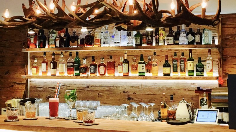 Eine Bar mit einer Holztheke, auf der verschiedene Getr&auml;nke stehen. Im Hintergrund sind Regale mit Spirituosen und eine Geweih-Lampe zu sehen., &copy; TOMAS