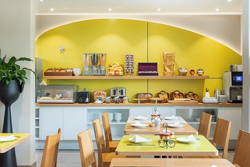 Modernes Frühstücksbuffet mit Brot, Obst, Cerealien und Kaffeemaschine. Helle, freundliche Atmosphäre mit Holztischen und grünen Akzenten., © adagio access Stuttgart Airport Messe