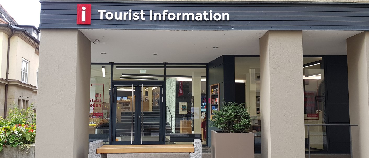 Eingang der Tourist Information Schwäbisch Hall mit Glasfront, Infomaterialien und einer Bank davor., © Leoni Friedrich Eingang der Tourist Information Schwäbisch Hall mit Glasfront, Infomaterialien und einer Bank davor., © Leoni Friedrich