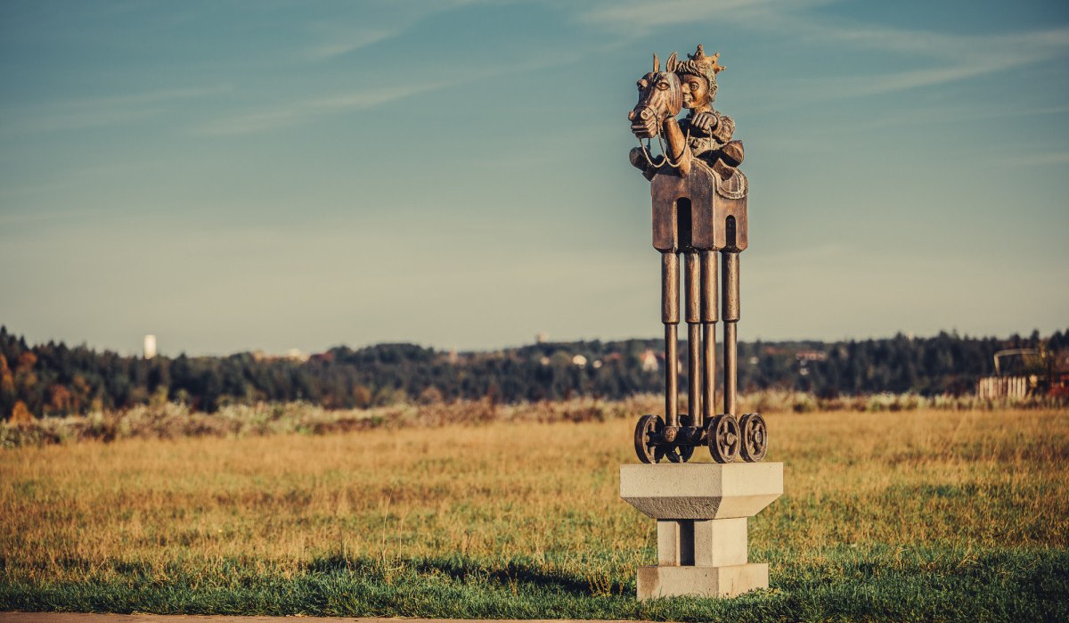 Eine Skulptur zeigt einen Reiter auf einem Pferd mit Rädern, platziert auf einem Sockel in einer weiten Wiesenlandschaft unter blauem Himmel., © Landratsamt Ostalbkreis