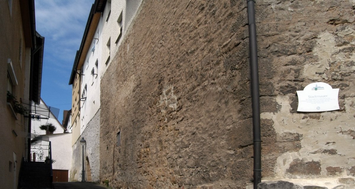 Schmale Gasse zwischen alten Geb&auml;uden, rechts eine historische Stadtmauer mit Schild. Blauer Himmel im Hintergrund., &copy; Natur.Nah. Sch&ouml;nbuch & Heckeng&auml;u