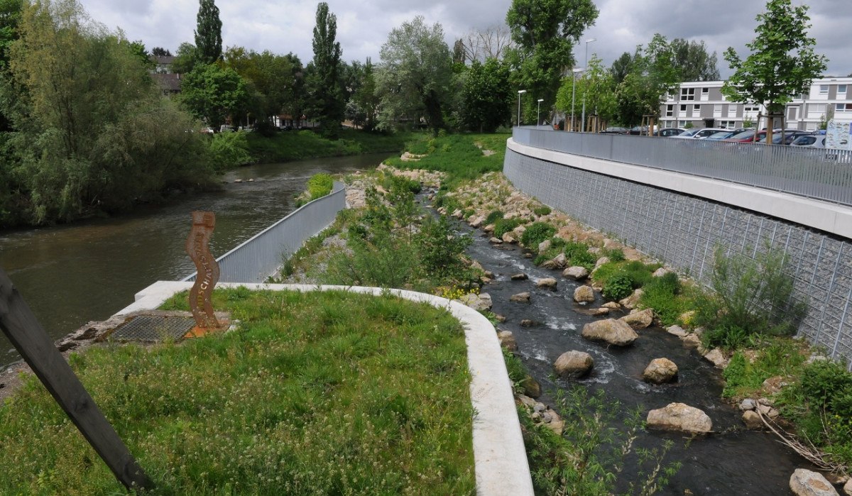 Flusslandschaft mit grünem Ufer, kleinem Bachlauf und Bäumen. Im Hintergrund sind Gebäude zu sehen., © Baumgärtner Flusslandschaft mit grünem Ufer, kleinem Bachlauf und Bäumen. Im Hintergrund sind Gebäude zu sehen., © Baumgärtner