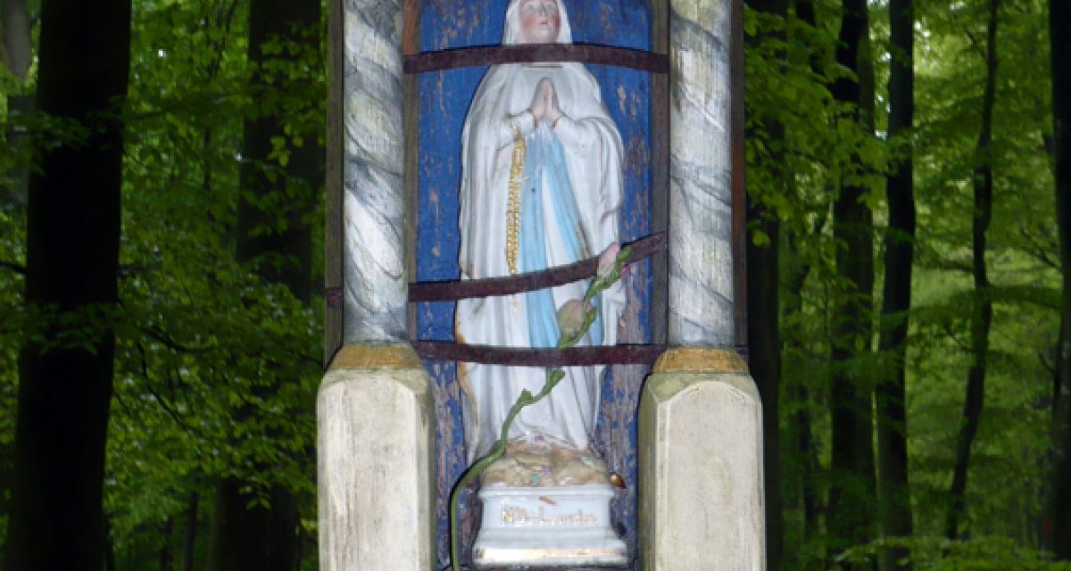 Eine Marienstatue in einem hölzernen Schrein, umgeben von Bäumen im Wald. Die Statue ist in einem gotischen Rahmen eingefasst. Eine Marienstatue in einem hölzernen Schrein, umgeben von Bäumen im Wald. Die Statue ist in einem gotischen Rahmen eingefasst.