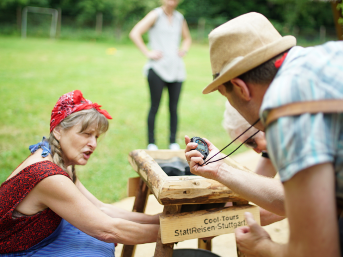 Eine Frau und ein Mann bei einer Outdoor-Aktivität. Der Mann hält eine Stoppuhr, die Frau arbeitet an einer Holzkonstruktion. Im Hintergrund eine weitere Person., © Cool-Tours StattReisen