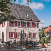 Ein Restaurant mit roten Fensterläden und Außensitzbereich auf einem gepflasterten Platz, umgeben von Bäumen und blauem Himmel., © Stadt Welzheim Ein Restaurant mit roten Fensterläden und Außensitzbereich auf einem gepflasterten Platz, umgeben von Bäumen und blauem Himmel., © Stadt Welzheim