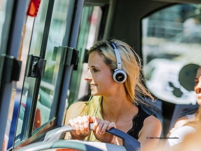 Eine Frau mit Kopfhörern sitzt in einem Bus und schaut lächelnd aus dem Fenster. Sonnenlicht fällt durch die Scheiben., © SMG, Thomas Niedermüller Eine Frau mit Kopfhörern sitzt in einem Bus und schaut lächelnd aus dem Fenster. Sonnenlicht fällt durch die Scheiben., © SMG, Thomas Niedermüller