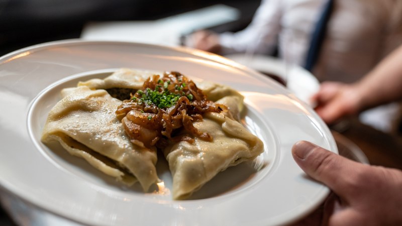 Ein Teller mit Maultaschen, garniert mit geschmolzenen Zwiebeln und Schnittlauch, wird serviert. Im Hintergrund ist eine Person unscharf zu sehen., © Stuttgart-Marketing GmbH, Martina Denker Ein Teller mit Maultaschen, garniert mit geschmolzenen Zwiebeln und Schnittlauch, wird serviert. Im Hintergrund ist eine Person unscharf zu sehen., © Stuttgart-Marketing GmbH, Martina Denker
