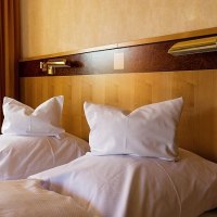 Zwei Betten mit weißen Kissen und Decken, Holzkopfteil und Leselampen in einem Gästezimmer., © Gästehaus Schlegel