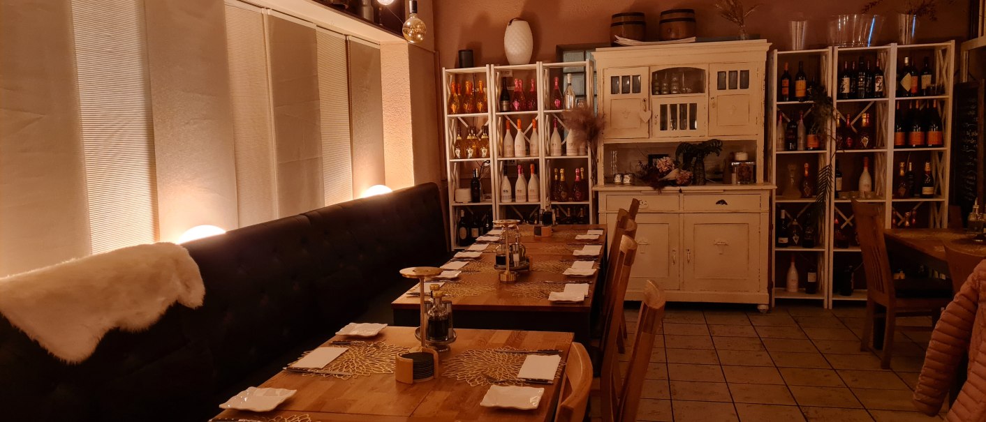 Ein gem&uuml;tlicher Restaurantinnenraum mit gedeckten Holztischen, dekorativen Platzsets und warmem Licht. Regale mit Flaschen und ein rustikaler Schrank erg&auml;nzen das Ambiente., &copy; RistoRocco