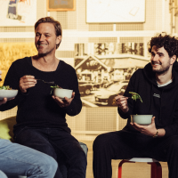 Drei Männer sitzen lachend zusammen und essen Salat in einem gemütlichen, künstlerisch gestalteten Raum., © Vhy! GmbH Drei Männer sitzen lachend zusammen und essen Salat in einem gemütlichen, künstlerisch gestalteten Raum., © Vhy! GmbH