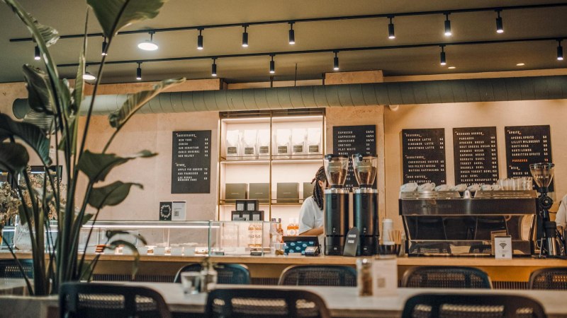 Modernes Café mit Theke, Kaffeemaschine und Speisekarten an der Wand. Pflanzen im Vordergrund, gemütliche Atmosphäre., © Maison Karl, Stuttgart Modernes Café mit Theke, Kaffeemaschine und Speisekarten an der Wand. Pflanzen im Vordergrund, gemütliche Atmosphäre., © Maison Karl, Stuttgart