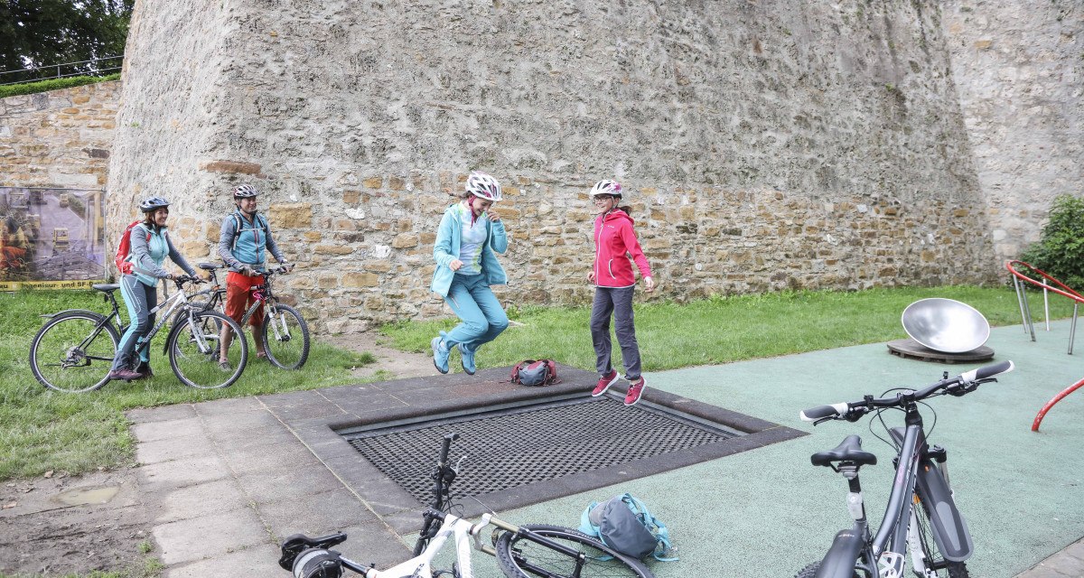Zwei Personen springen auf einem Trampolin neben Fahrrädern vor einer alten Steinmauer. Weitere Personen stehen mit Fahrrädern daneben., © Schwäbische Alb Tourismusverband e.V.