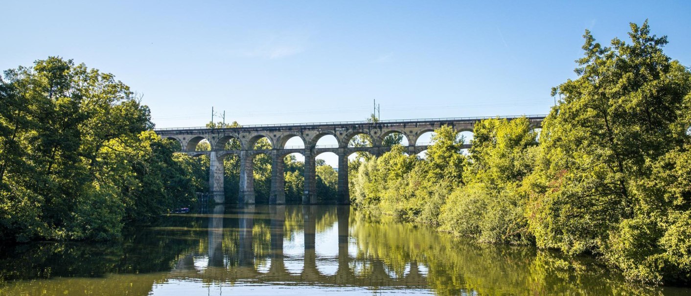 Ein Viadukt überspannt einen Fluss, umgeben von grünen Bäumen und klarem Himmel. Die Brücke spiegelt sich im Wasser wider., © SMG, Sarah Schmid Ein Viadukt überspannt einen Fluss, umgeben von grünen Bäumen und klarem Himmel. Die Brücke spiegelt sich im Wasser wider., © SMG, Sarah Schmid