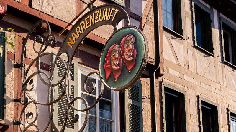Ein kunstvoll gestaltetes Schild der Narrenzunft h&auml;ngt an einem Fachwerkhaus in Weil der Stadt. Es zeigt zwei Masken mit roten Kopft&uuml;chern., &copy; Olivia + RichiPhotoart
