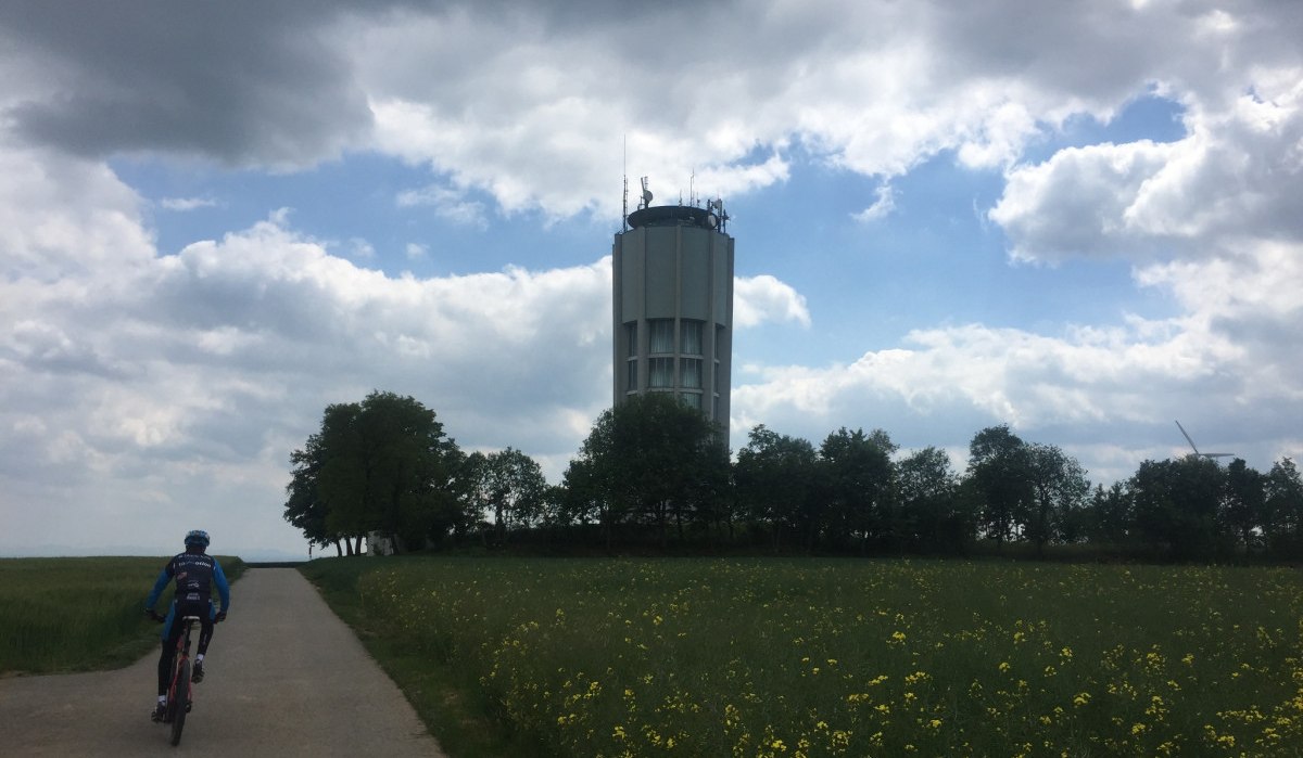 Ein Radfahrer auf einem Weg zum Wasserturm beim Eisberg Jettingen, umgeben von Bäumen und blühenden Wiesen unter bewölktem Himmel., © www.pro-cycl.de Ein Radfahrer auf einem Weg zum Wasserturm beim Eisberg Jettingen, umgeben von Bäumen und blühenden Wiesen unter bewölktem Himmel., © www.pro-cycl.de