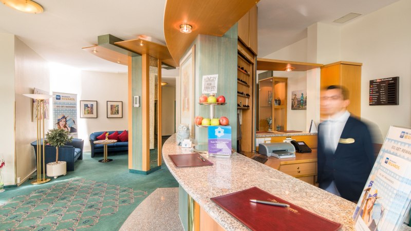 Hotelrezeption mit geschwungener Theke, Dekoration mit Äpfeln und Eule, sowie einem verschwommenen Mitarbeiter im Anzug. Sitzbereich im Hintergrund., © Plaza Hotel Blankenburg Ditzingen