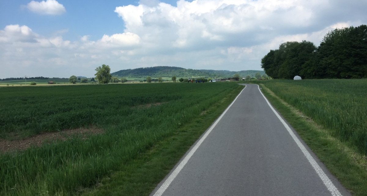Ein asphaltierter Radweg schlängelt sich durch eine grüne Landschaft mit Feldern und Bäumen. Der Himmel ist blau mit einigen Wolken., © www.pro-cycl.de Ein asphaltierter Radweg schlängelt sich durch eine grüne Landschaft mit Feldern und Bäumen. Der Himmel ist blau mit einigen Wolken., © www.pro-cycl.de