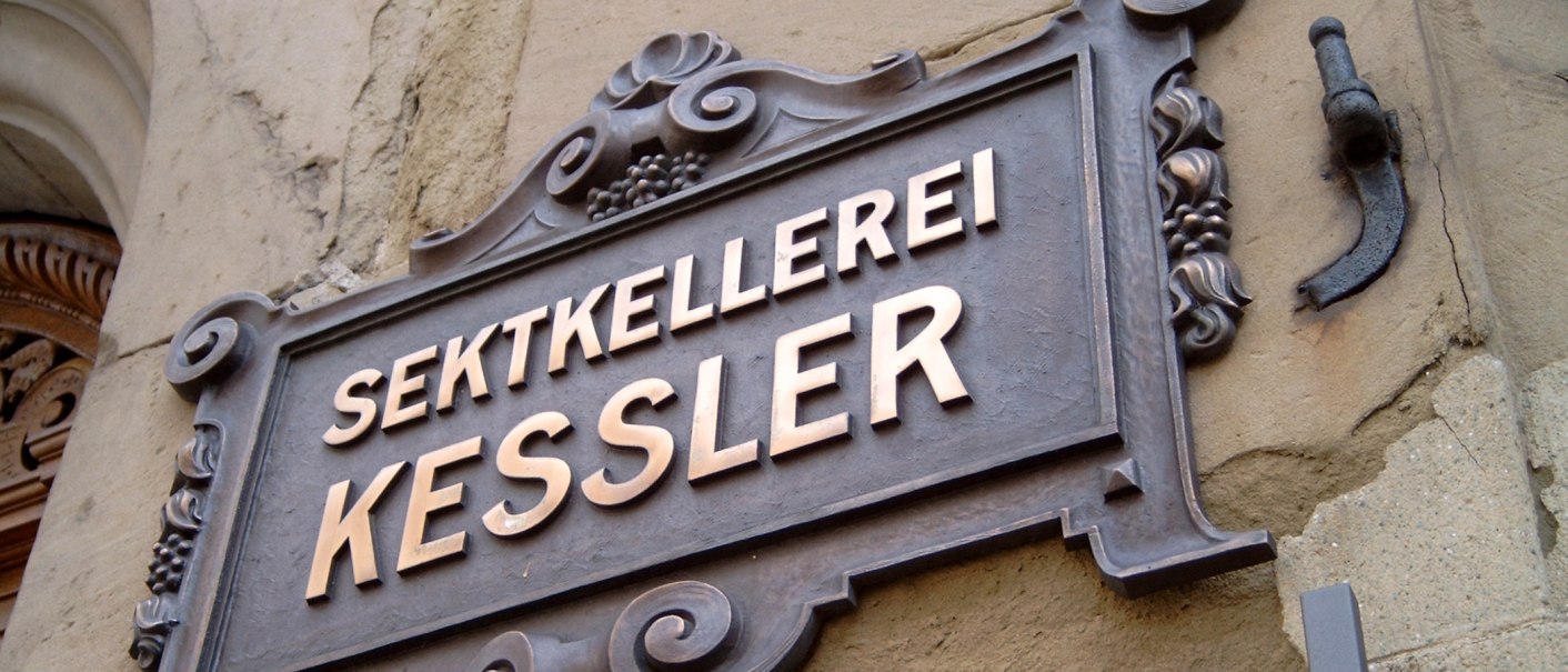 Ein kunstvoll gestaltetes Metallschild mit der Aufschrift 'Sektkellerei Kessler' an einer historischen Steinwand., © Kessler Sekt