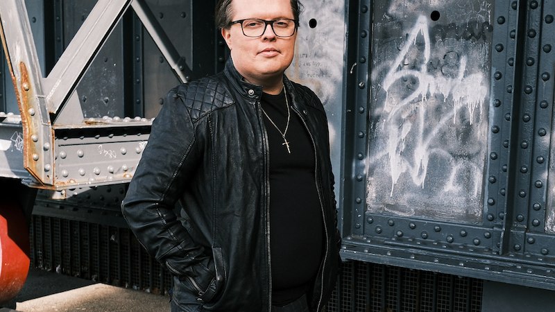 Person in schwarzer Lederjacke und Brille steht vor einer metallischen Wand mit Graffiti., © Scala Kultur Live gGmbH