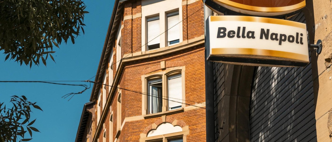 Ein Schild mit der Aufschrift 'Bella Napoli' hängt an einem Backsteingebäude. Der Himmel ist klar und blau, ein Baum ist im Vordergrund sichtbar., © SMG, Sarah Schmid