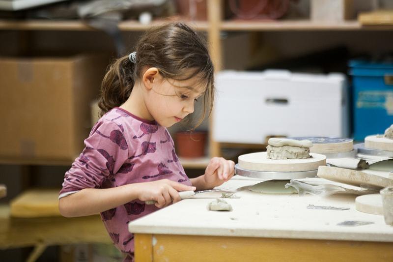 Ein Kind in einem rosa Oberteil formt konzentriert Ton auf einem Tisch in einem Workshop. Im Hintergrund sind Regale mit Materialien zu sehen., © Kunstschule Labyrinth Ein Kind in einem rosa Oberteil formt konzentriert Ton auf einem Tisch in einem Workshop. Im Hintergrund sind Regale mit Materialien zu sehen., © Kunstschule Labyrinth