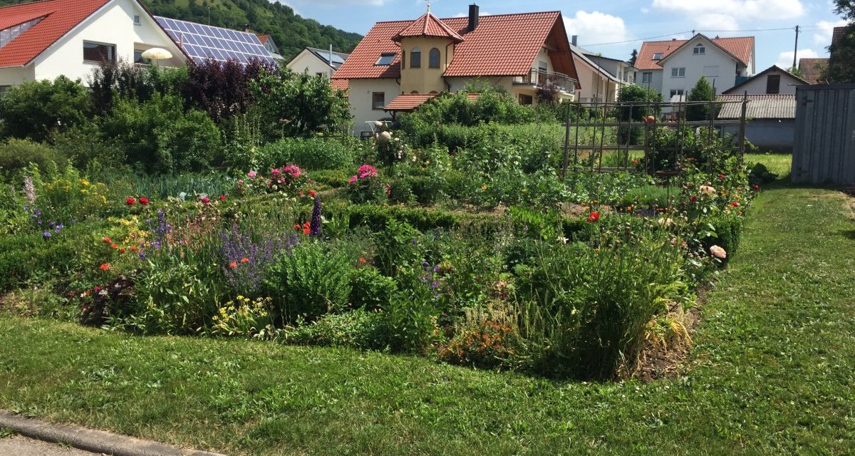 Ein bl&uuml;hender Vorgarten mit bunten Blumen und Str&auml;uchern vor mehreren H&auml;usern mit roten D&auml;chern und Solarpanelen. Im Hintergrund sind gr&uuml;ne H&uuml;gel zu sehen., &copy; www.pro-cycl.de